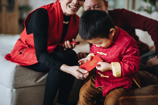 Cute little grandson dressed in red traditional Chinese costume receives red envelops (lai see) with both hands from grandparents joyfully in Chinese New Year