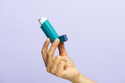 Female hand holding blue asthma inhaler on the purple background.