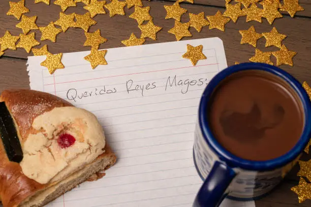 Three wisemen letter that says: Dear wisemen in Spanish with golden stars, chocolate and a slice of traditional Epiphany cake on wooden table