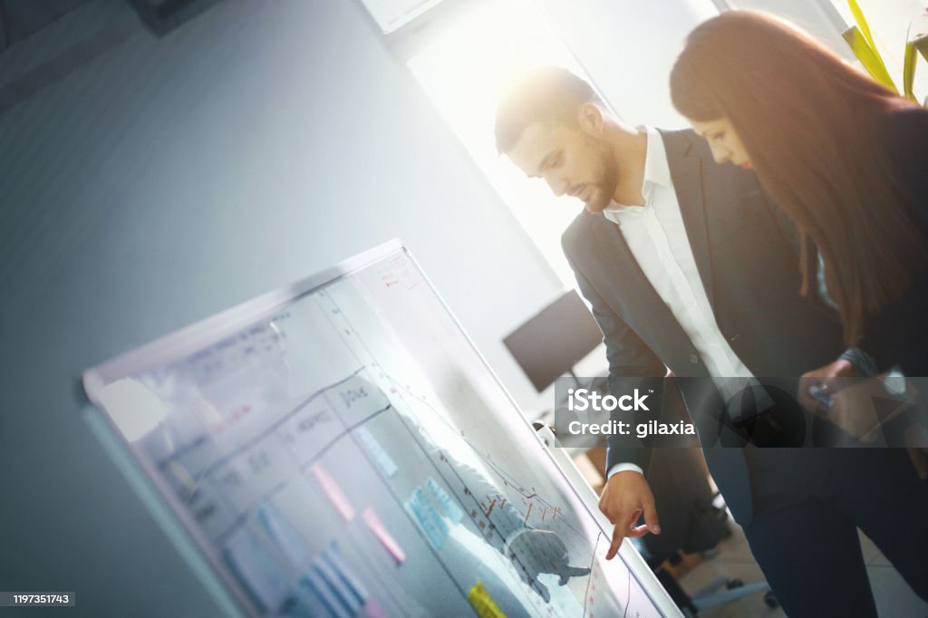 Entrepreneur evaluation meeting. Closeup side view of two late 20's entrepreneurs analyzing data and business figures by looking at a white board. Graphs and numbers are plummeting in the last quarter. Performance Review Stock Photo