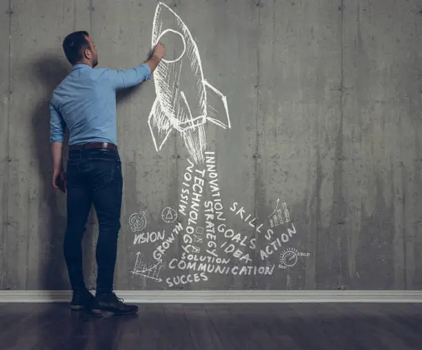 Businessman drawing a rocket with business words as engine smoke on a concrete wall.