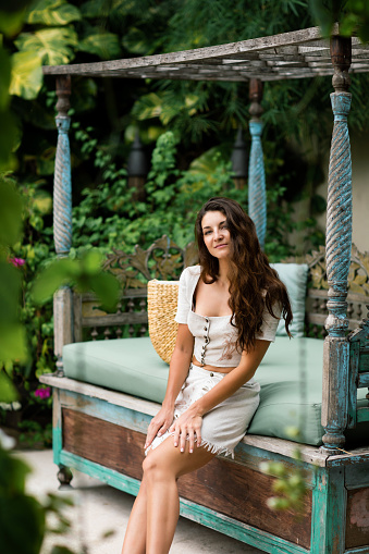 Beautiful brunette woman with long hair in linen top and skirt with straw handbag, posing in tropical background on Bali. Natural outfit and organic summer style. Fashion trend.