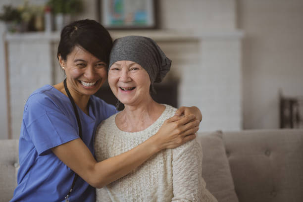 Nurse Hugging Her Cancer Patient stock photo A young Cambodian female nurse wraps her arms around her elderly cancer patients shoulders as they smile and share an embrace.  The elderly woman is dressed casually in a sweater and head scarf and is wearing glasses, while the nurse is wearing blue scrubs. cambodian ethnicity stock pictures, royalty-free photos & images
