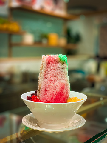 Ice Kacang, an icy dessert of shave ice with toppings of red beans, agar, cendol jelly, fruits and syrups. Popular in Singapore and Malaysia. The dessert is plated in a white, plain ceramic bowl with an underliner; placed on a serving counter in the dessert market stall. In the background is the back counter of the stall.