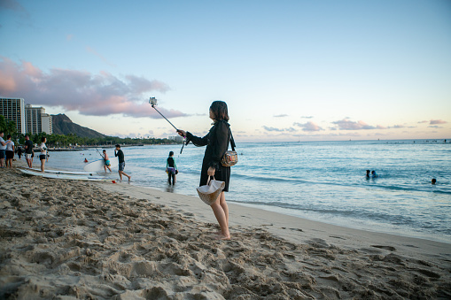 02/04/2019- Asian woman taking selfie in Honolulu using selfie stick and mobile phone on Waikiki beach. Tourists along the coastline, Oahu Island, Hawaii, USA