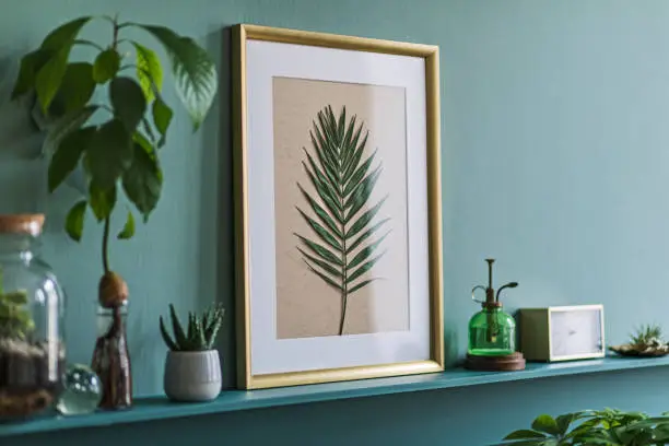 Photo of Interior design of living room with gold mock up photo frame on the green shelf with beautiful plants, cacti in different hipster and design pots. Elegant personal accessories. Home jungle. Template.