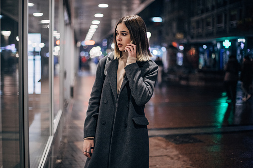 Beautiful young woman walking in the city downtown at night and using mobile phone while window shopping