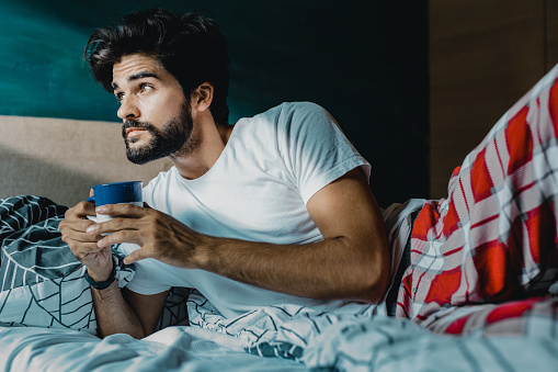 Young man is in the bed in the morning, he is looking at the window while holding coffee cup