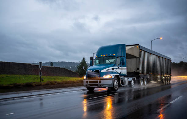 Big rig bonnet blue semi truck transporting cargo in covered bulk semi trailer running on the wet glossy road with raining weather evening Big rig day cab bonnet blue semi truck with roof spoiler transporting commercial cargo in covered corrugated bulk semi trailer running on the evening wet glossy slippery road with raining weather truck rain stock pictures, royalty-free photos & images