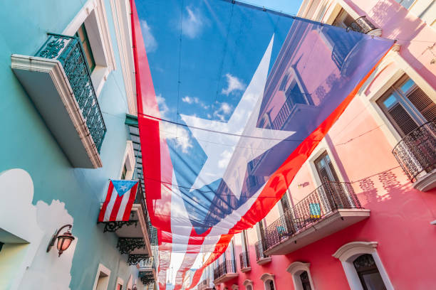 Large flag of Puerto Rico above the street in the city center of San Juan. Colorful image of city centre of San Juan with large Puerto Rican flag above the street. Blue and pink buildings in the street. Sunny day. Red and white stripes, white star and blue colored national flag of Puerto Rico. puerto rico photos stock pictures, royalty-free photos & images