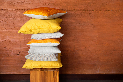 Stock of colorful pillows on wood chair against textured terracotta wall. Pile of soft yellow and orange cushions on stool near wall with space for text. Modern and cozy home concept.