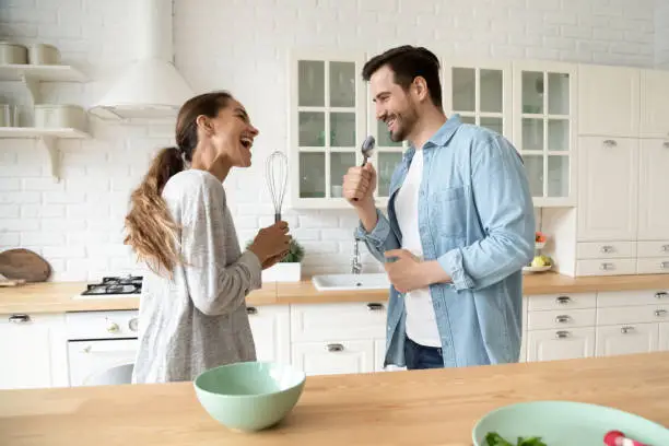 Funny young couple singing in kitchenware microphones in kitchen, happy carefree husband and wife hold beater spoon together having fun preparing dinner breakfast cooking dancing listen music at home