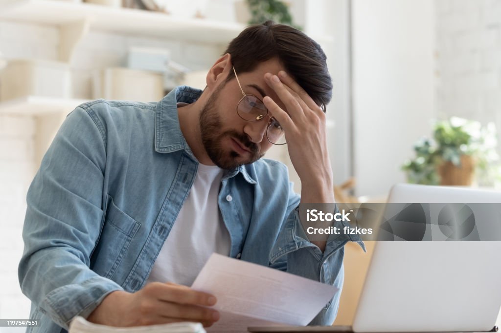 Upset frustrated young man holding reading postal mail letter Upset frustrated young man reading bad news in postal mail letter paper document sit at home table, depressed stressed guy worried about high bill tax invoice, overdue debt notification money problem Financial Bill Stock Photo