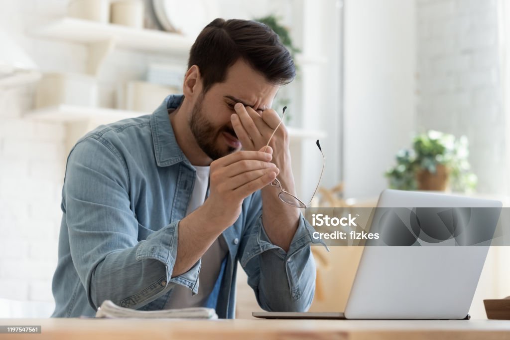 Tired young man feel eyestrain holding glasses fatigued from computer Tired young man feel pain eyestrain holding glasses rubbing dry irritated eyes fatigued from computer work, stressed man suffer from headache bad vision sight problem sit at home table using laptop Men Stock Photo