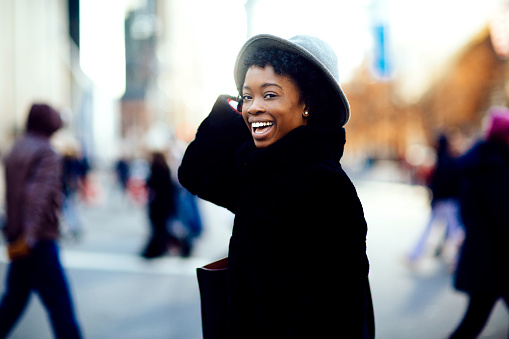 Portrait of a happy smiling woman in faux fur coat and hat laughing at camera while crossing a busy urban street