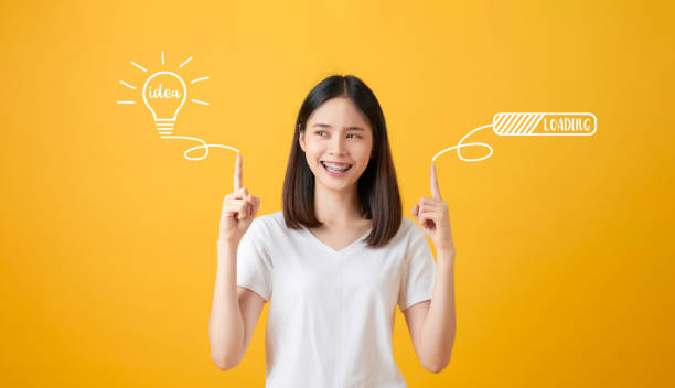 istock Young smiling Asian woman pointing hands to light bulb with text ideas and loading brainstorm on yellow background. 1197583693