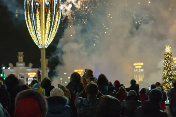 Defocused photography of bright fireworks in New Year's night on city street. Celebration of New Year 2020. VDNkH sky in mid night. Backs of people. Holidays and lifestyles concepts. Rear / backs view