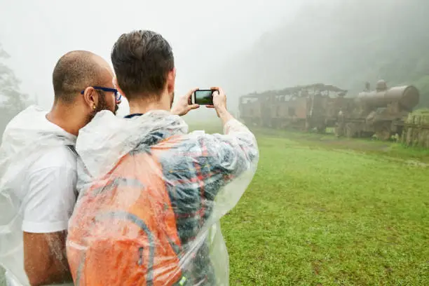 Rear view shot of two young male friends in raincoat taking pictures of a rusty train on a rainy day with mobile phone camera