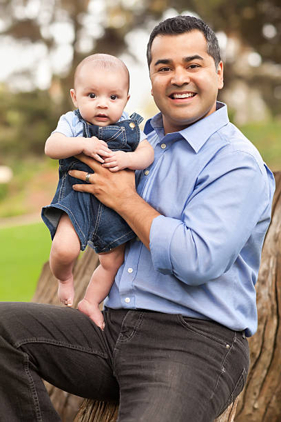 A happy latin father staning outdoors with a baby and a little boy, looking at the camera and smiling.