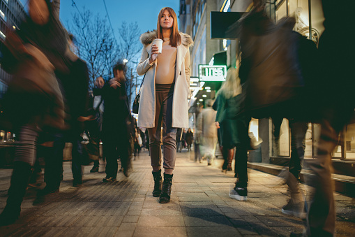 Beautiful young pregnant woman walking over sidewalk in city at night and she is surrounded with other people blurred in motion. She wears elegant warm clothes and holding coffee to go in her hand.