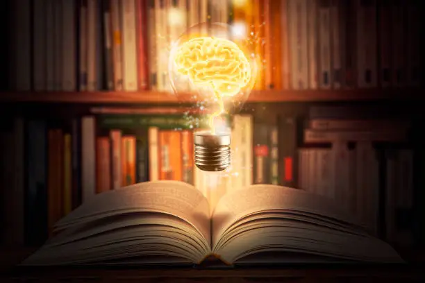 Glowing and shining brain lightbulb over an open book with a bookshelf as background. Knowledge, study, cognition, learning, literacy, library 3d illustration concepts. Mixed media.