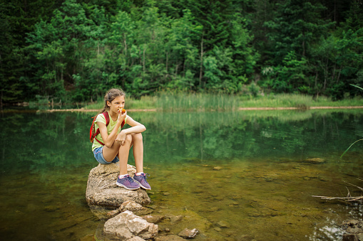 Girl sitting on the rock in the river, enjoying nature and eating apple. She is very cute and has healthy habits