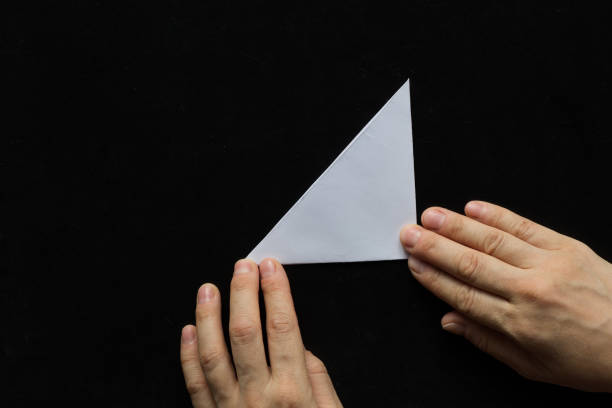 istock Making snowflake. Step 2. Woman hands holding paper folded small triangle on the black background 1197676038