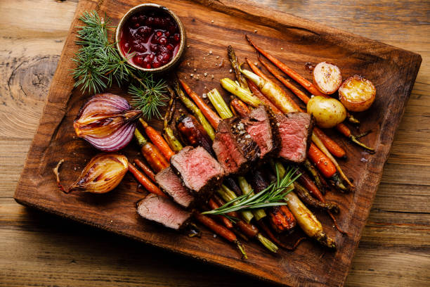 grilled sliced venison steak with baked vegetables and berry sauce on wooden background - venison imagens e fotografias de stock