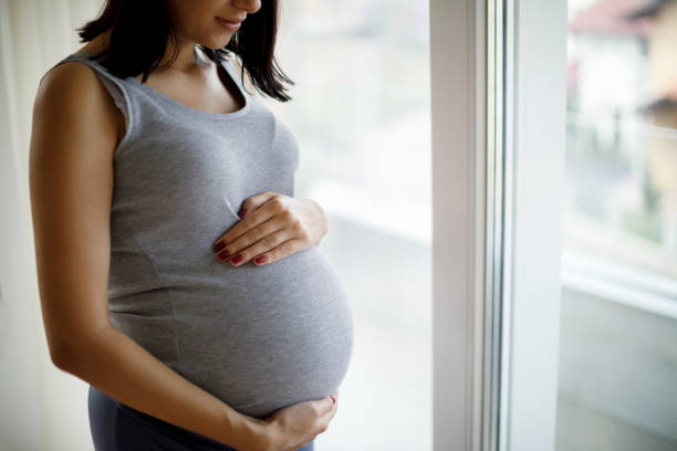 verticale de jeune femme enceinte restant par la fenêtre - holding belly photos et images de collection
