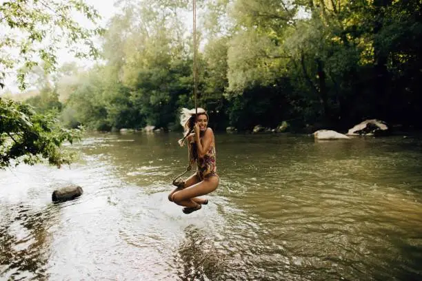 Photo of a young woman who is spending hot summer day on the river, swinging on a rope that looks like liana