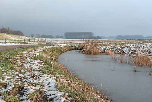 Partially snowy Dutch landscape with a curved stream and country road. In the background is a dyke. There is a thin layer of ice on the water. The sky is gray, more snow is coming soon.