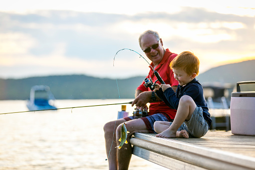 A grandfather is teaching his grandson to fish during sunset in summer. They are both sitting on the dock and are concentrated on their activity. The little redhead boy caught a fish and he is smiling and very happy. It is a beautiful summer day. Across the lake, there is a mountain.
