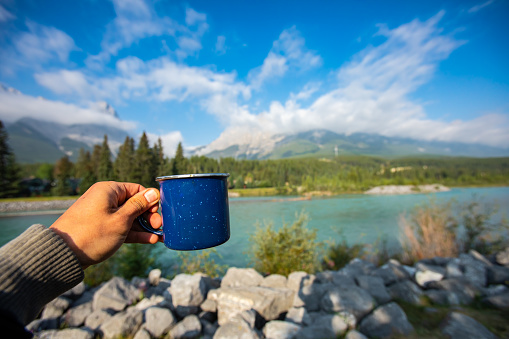 Selective focus of a persons hand holding a blue cup of tea or coffee with in the background a clear lake with trees and mountains behind it