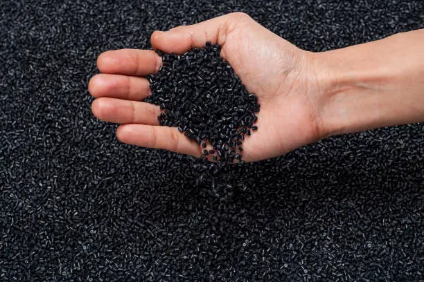 CLOSE-UP FLAT LAY OF BLACK POLYMER GRANULES ON GLASS TABLE WITH HUMAN HAND HOLDING, CHECKING THE GRANULES AND DROPPING BACK ON TABLE