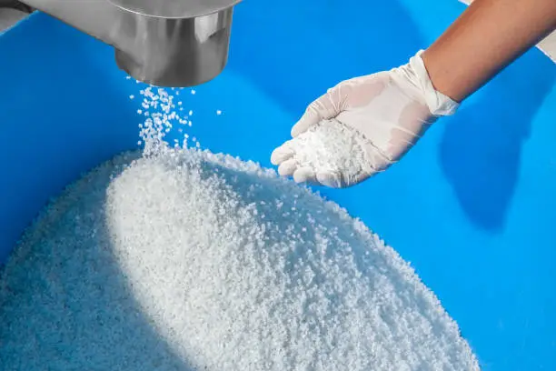 FINAL MANUFACTURED WHITE POLYMER GRANULES BEING ACCUMULATED IN BLUE PIT, HUMAN HAND WEARING WHITE GLOVE WITH SOME GRANULES ON PALM, CHECKING BEFORE FINAL PACKAGING.