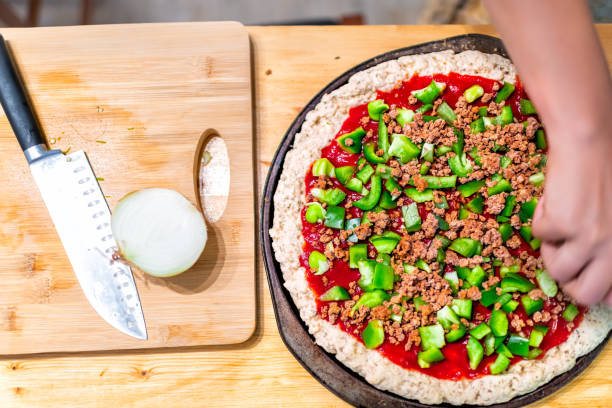 Flat top view down of raw pizza dough crust before baking made with gluten free flour and woman hands adding chopped green bell peppers on tomato sauce Flat top view down of raw pizza dough crust before baking made with gluten free flour and woman hands adding chopped green bell peppers on tomato sauce pizza topping stock pictures, royalty-free photos & images