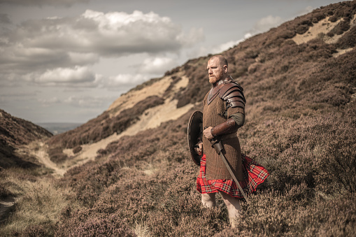 A lone redhead individual Scottish viking sword wielding warrior man wearing a kilt on highland moors