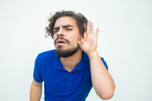 Serious focused guy listening to secret or gossip with hand at ear. Handsome bearded young man in blue casual t-shirt posing isolated over white background. Hearing concept