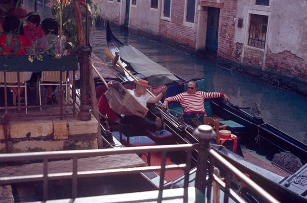 Venice, Venzien, Italy, 1984. Two gondoliers are waiting for passengers in their gondolas. Also: restaurant with guests, buildings and a canal.