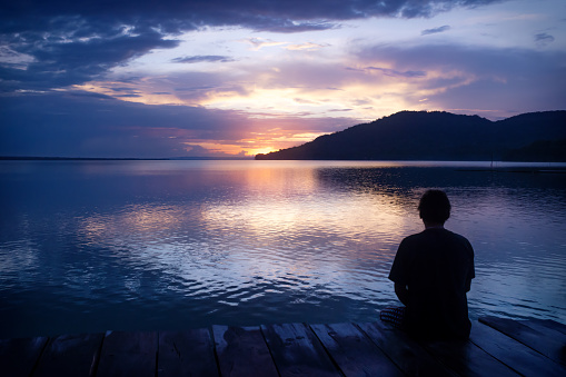 El Remate, Peten, Guatemala - 20 September 2019: Man sitting on dock with orange sunset on dramatic purple blue sky along lake Itza