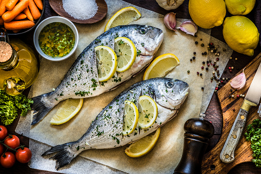 Top view of two seasoned raw sea breams on a wax paper surrounded by garlic, parsley, lemon, salt and pepper for seasoning the fish, some carrots, cherry tomatoes and a cutting board with a kitchen knife on top. Low key DSLR photo taken with Canon EOS 6D Mark II and Canon EF 24-105 mm f/4L