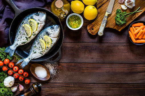Top view of two seasoned raw sea breams on a baking pan surrounded by garlic, parsley, lemon, salt and pepper for seasoning the fish and some carrots and cherry tomatoes. The objects are at the top and at the left side of the image leaving a useful copy space at the lower right corner on the rustic wooden backdrop. Low key DSLR photo taken with Canon EOS 6D Mark II and Canon EF 24-105 mm f/4L