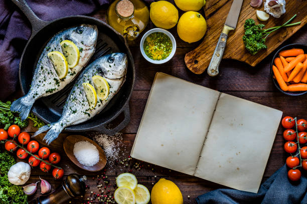 Top view of two seasoned raw sea breams on a baking pan surrounded by garlic, parsley, lemon, salt and pepper for seasoning the fish and some carrots and cherry tomatoes. At the center of the image is an opened cookbook with copy space ideal for a recipe, a text or a logo. The objects are on a rustic wooden backdrop. Low key DSLR photo taken with Canon EOS 6D Mark II and Canon EF 24-105 mm f/4L