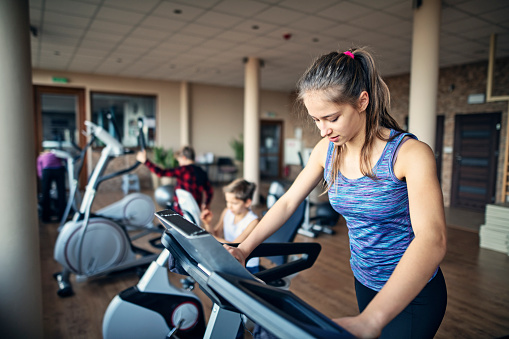 Teenage girl training in exercise room. The girl is using a cross trainer. Her brothers are exercising in the background.\nNikon D850
