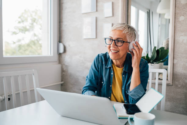 Mature woman freelancing from her home office Mature woman with headphones working online from her home. podcasting photos stock pictures, royalty-free photos & images