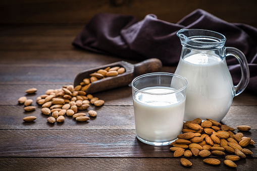 Front view of a jug and a drinking glass full of almond milk. The glass and the jug are at the right side of the image on a rustic wooden table, next to the glass its a heap of almonds. This image is a part of a vegan milk series. Low key DSLR photo taken with Canon EOS 6D Mark II and Canon EF 24-105 mm f/4L