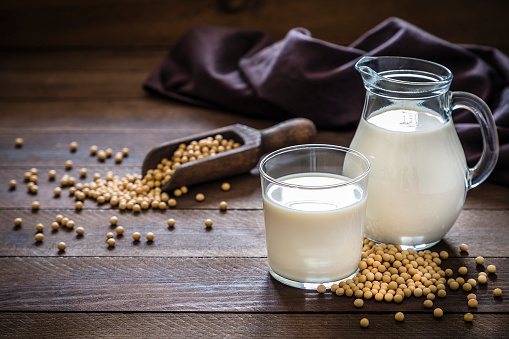 Front view of a jug and a drinking glass full of soy milk. The glass and the jug are at the right side of the image on a rustic wooden table, next to the glass its a heap of soy beans. This image is a part of a vegan milk series. Low key DSLR photo taken with Canon EOS 6D Mark II and Canon EF 24-105 mm f/4L