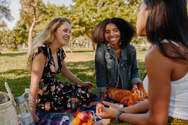 gruppe von lächelnden multiracial weiblichen besten freundinnen sitzen zusammen auf decke mit früchten genießen beim picknick im park - gruppe von gesunden freunden bei einem picknick - picknick stock-fotos und bilder
