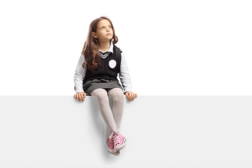 Schoolgirl in a uniform sitting on a blank panel and looking up isolated on white background