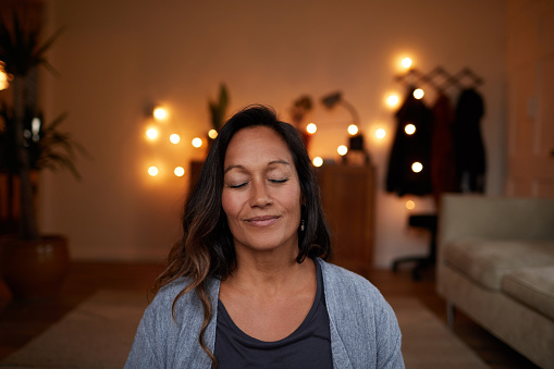 Serene woman smiling with her eyes closed while sitting on her living room floor practicing yoga